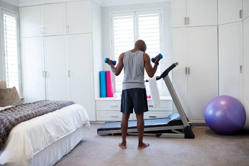 Rear view of man exercising with dumbbells in bedroom at home.