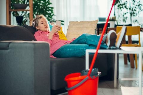 Woman lounging on a couch, absorbed in her smartphone, with cleaning supplies nearby.