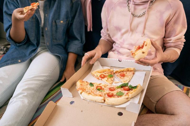 Two relaxed teenagers in casualwear eating pizza from box while sitting on the floor.