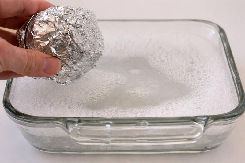 Ai rendering of a glass casserole dish filled with soapy water. A person off-screen is holding a ball of crumpled up aluminum foil over the scene