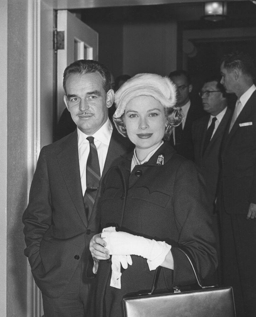 Prince Rainier and Grace Kelly standing in a doorway. A group of men stand together in a group behind them