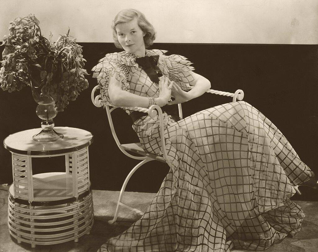 Katharine Hepburn sitting in a chair, next to a small table with a plant atop it