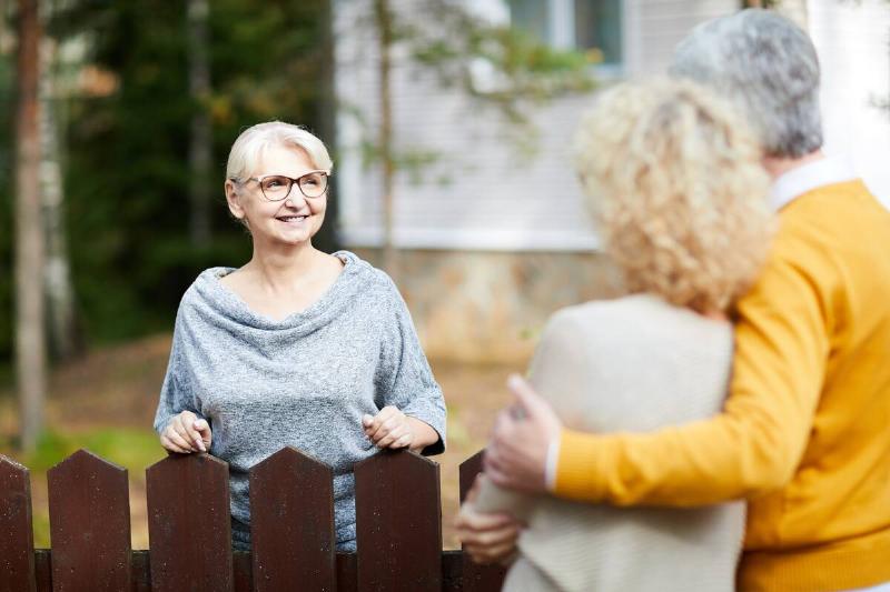 Blonde woman in eyeglasses and grey pullover looking at her neighbors during talk through fence.