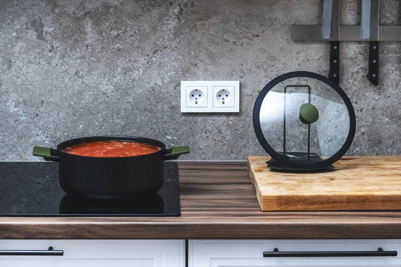 A glass pot lid on a cutting board beside a glass stovetop with a pot cooking on it. 