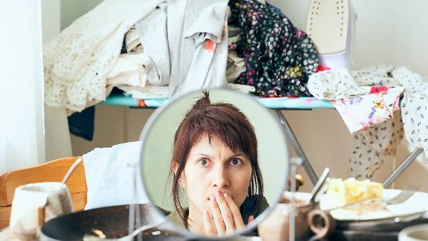 Woman looking at her reflection in front of a pile of messy clothes and dishes. 