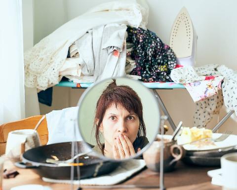 Woman looking at her reflection in front of a pile of messy clothes and dishes. 