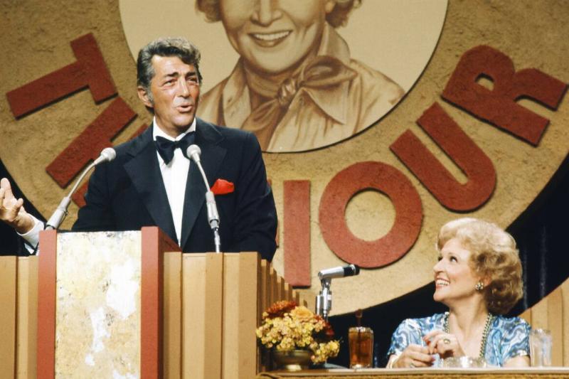 Dean Martin speaking at a podium, while Betty White looks at him from a table nearby