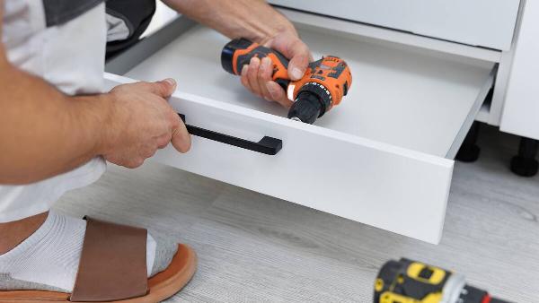 Person crouching to use a power drill to install a new handle on a drawer. 