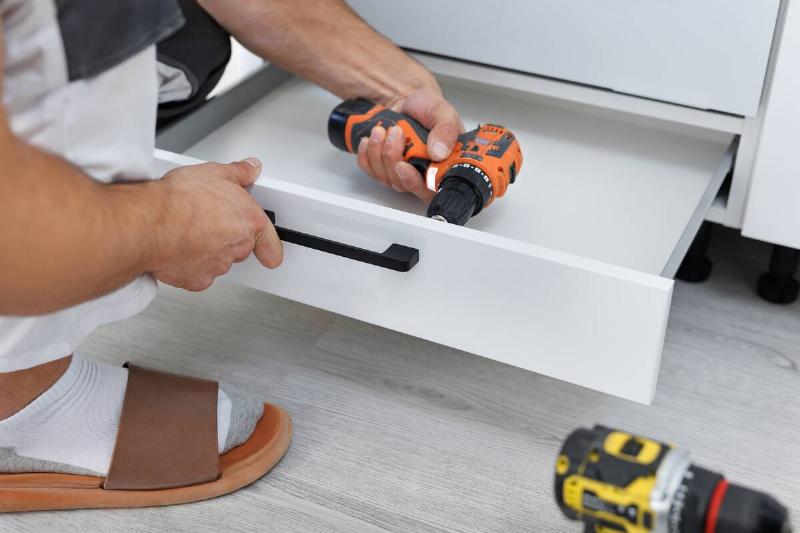 Person crouching to use a power drill to install a new handle on a drawer. 