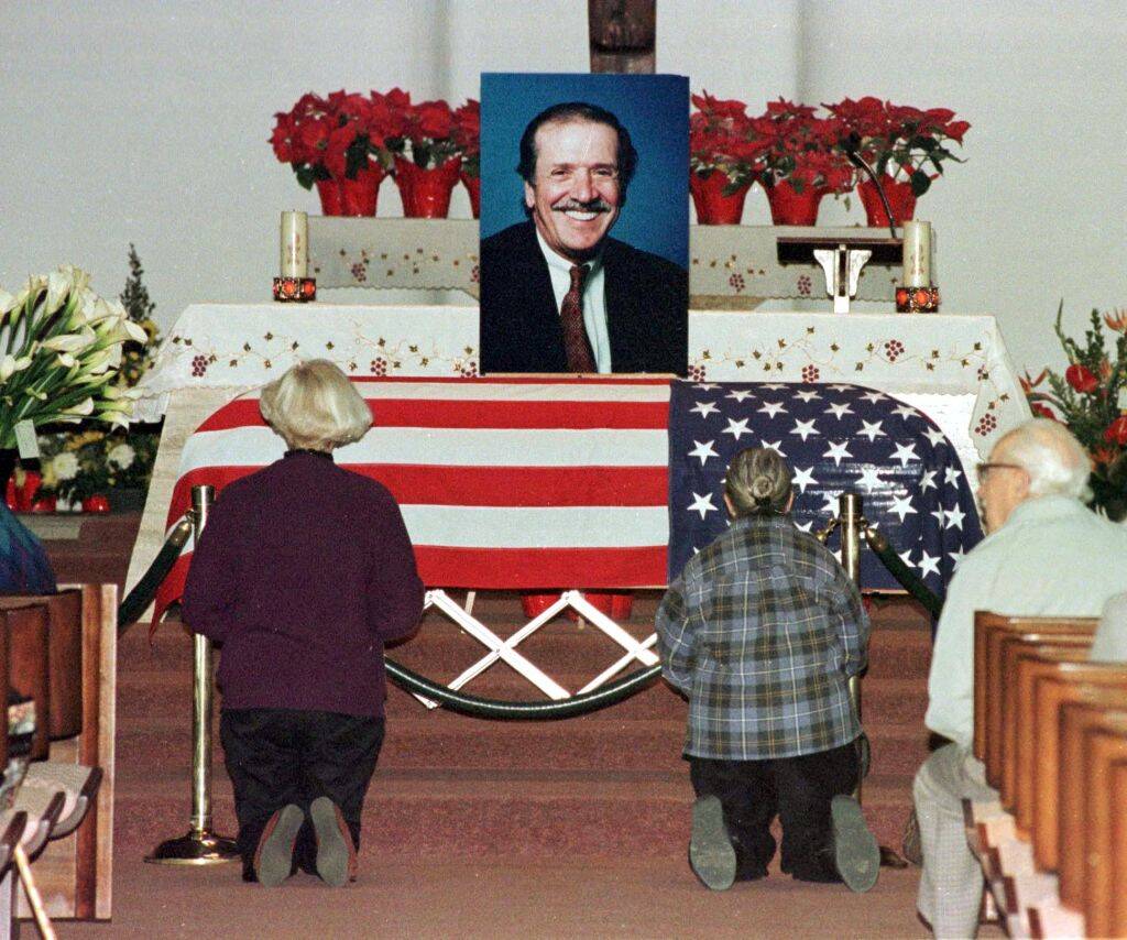Two individuals kneeling before a US flag-draped coffin that has a picture of Sonny Bono atop it