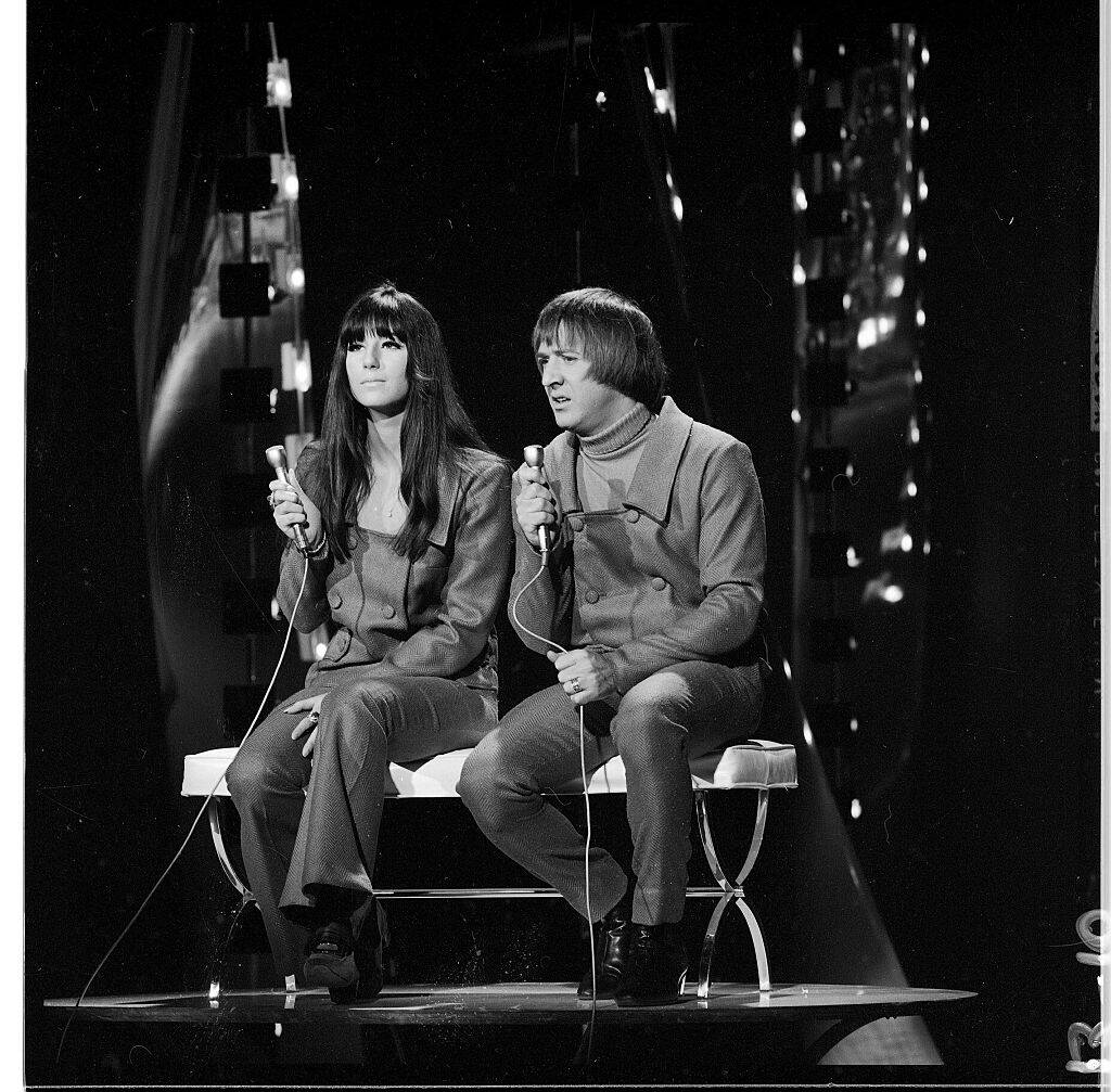 Sonny Bono and Cher sitting together on stage, each holding a microphone