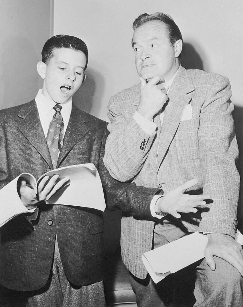 Tony Hope looking down at a script while Bob Hope stands beside him, looking intrigued