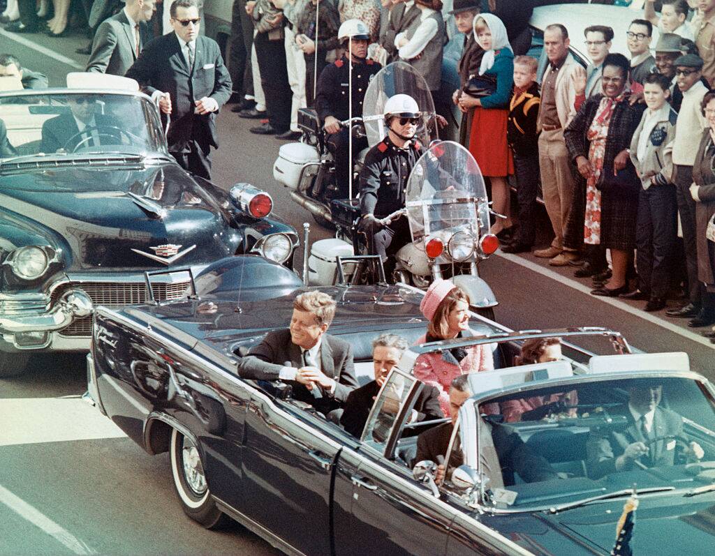 Jackie Kennedy, John F. Kennedy and John Connally riding in a motorcade. A crowd stands around them, and a police officer rides alongside them on a motorcycle