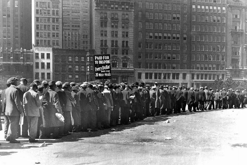 Men lined up alongside a row of building in New York City, New York. A sign is held above them, which reads, 