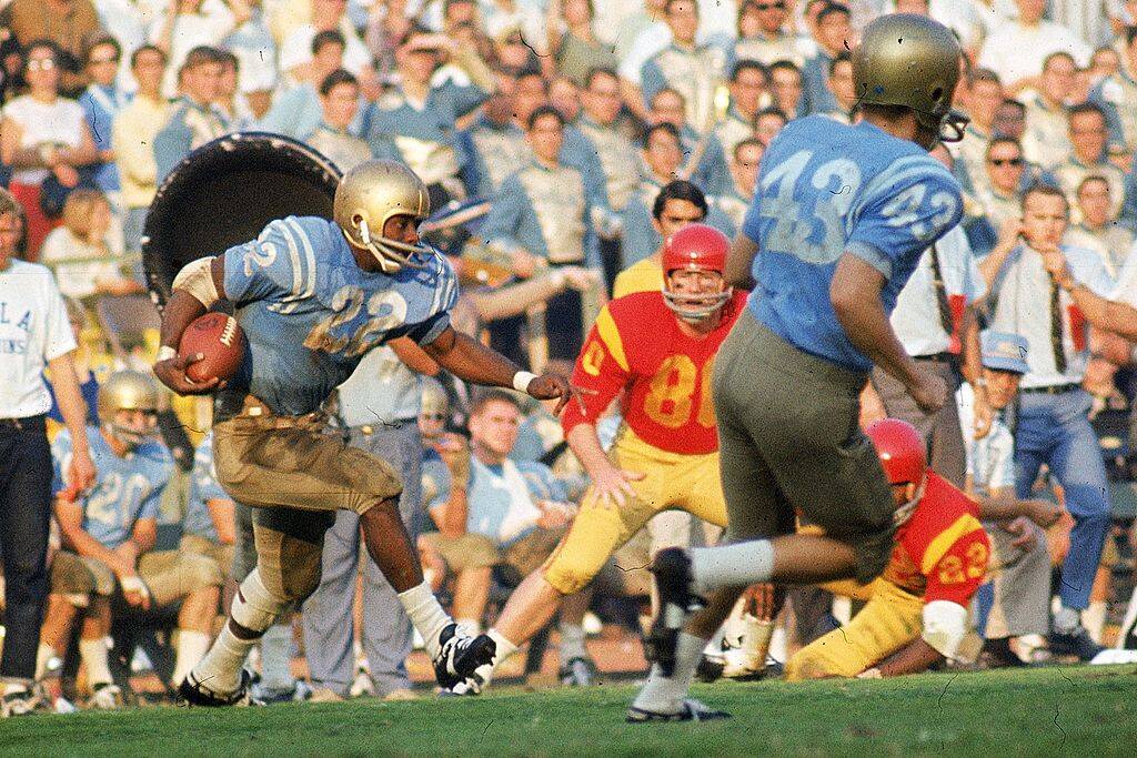 Football players running on a pitch. One has a football under his arm. A large crowd watches from the stands