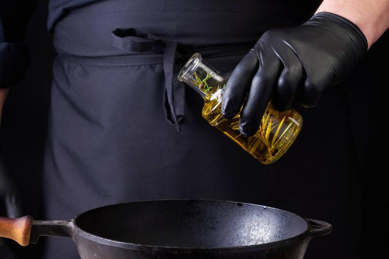 шеф-повар в черных латексных перчатках наливает оливковое масло из-2026-01-11-08-15-11-utc Человек наливает масло в чугунную кастрюлю.