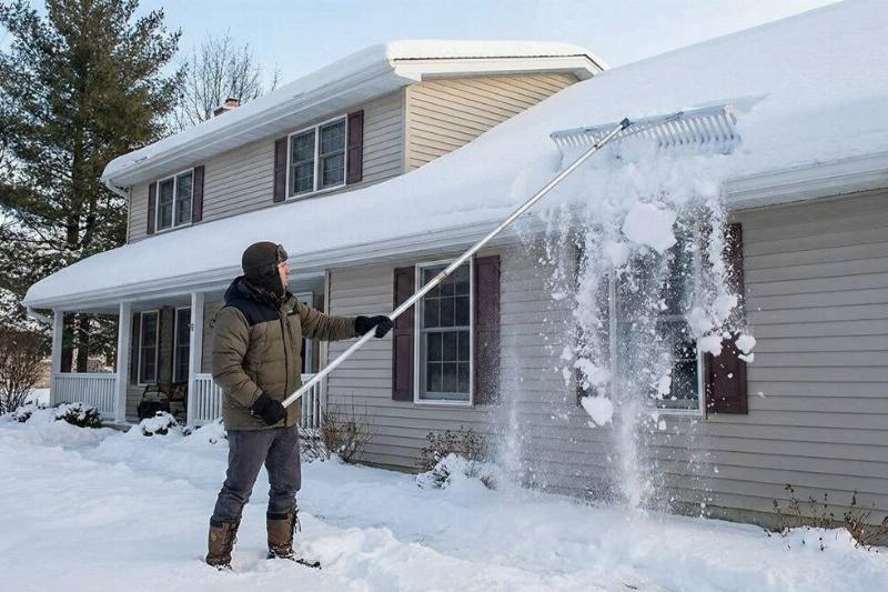 A man cleaning snow off the roof of his house using a roof rake. 