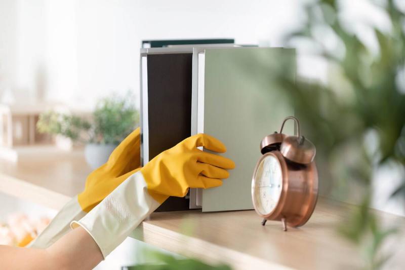 Close-up of hands in yellow gloves placing books on shelf with clock on it. 