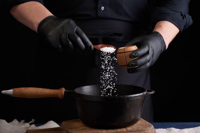 мужчина-повар в черной униформе и латексных перчатках-соль-f-2026-01-11-08-16-03-utc Мужчина кладет соль в чугунный горшок.