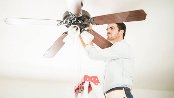 A man standing on a ladder to change the direction that a ceiling fan is spinning. 
