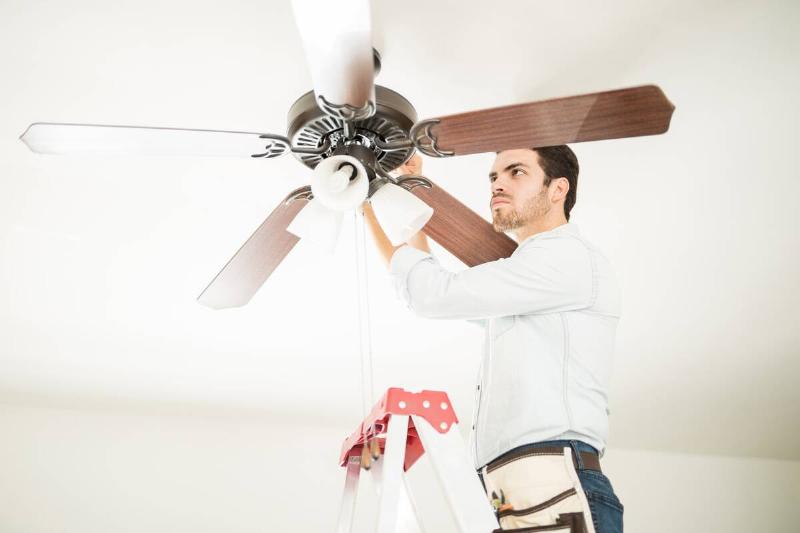 A man standing on a ladder to change the direction that a ceiling fan is spinning. 