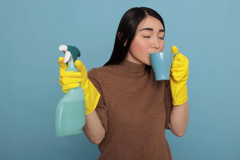 Woman drinking coffee from a mug while holding a spray bottle and wearing rubber gloves. 