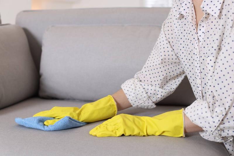 Woman in gloves cleaning sofa with rag at home.