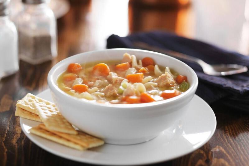 Bowl of chicken noodle soup on a table with utensils and salt and pepper shakers. Crackers are placed on the plate that the bowl is atop of