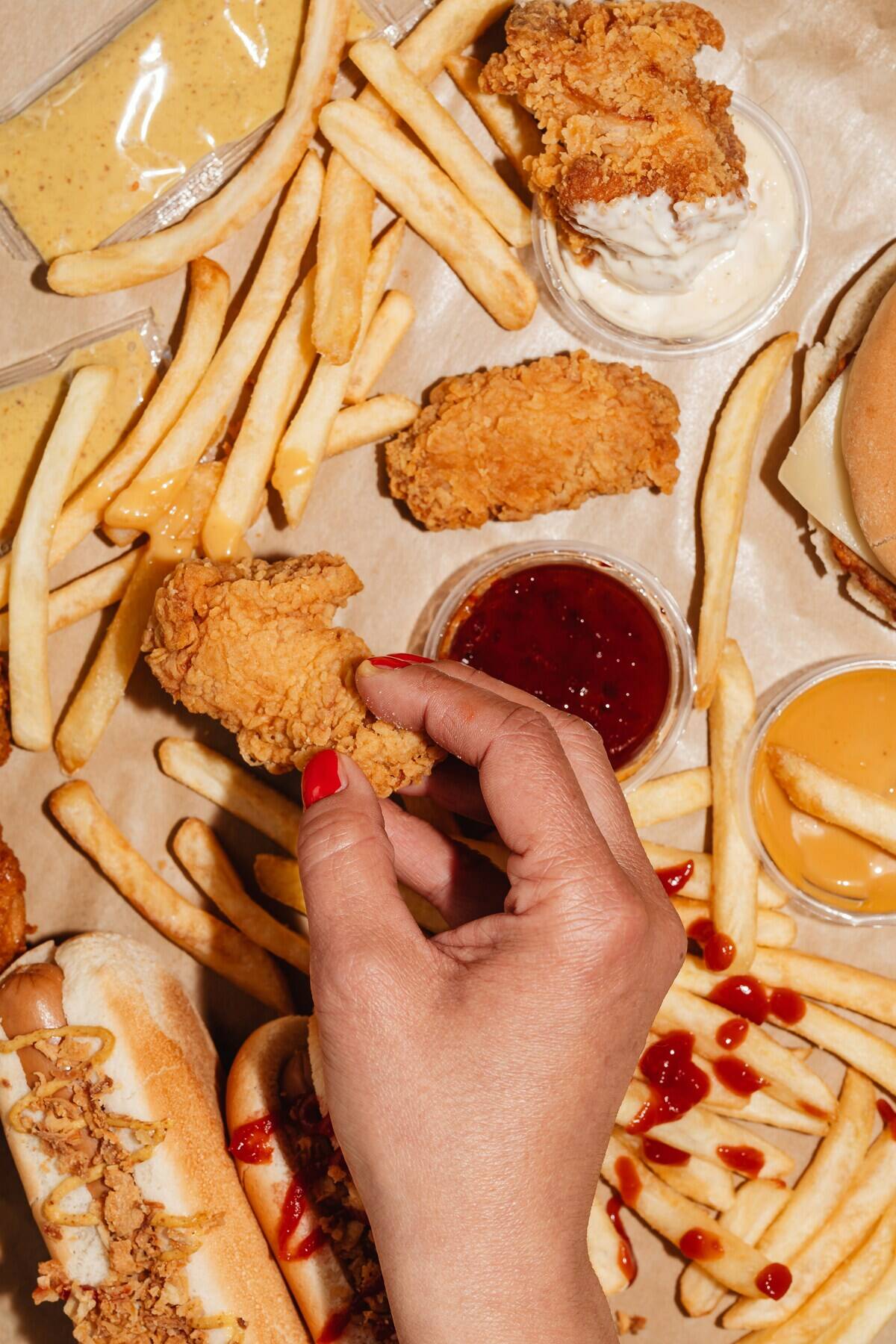 Person off-screen holding a breaded chicken wing above a table covered in fries, condiments and a hot dog