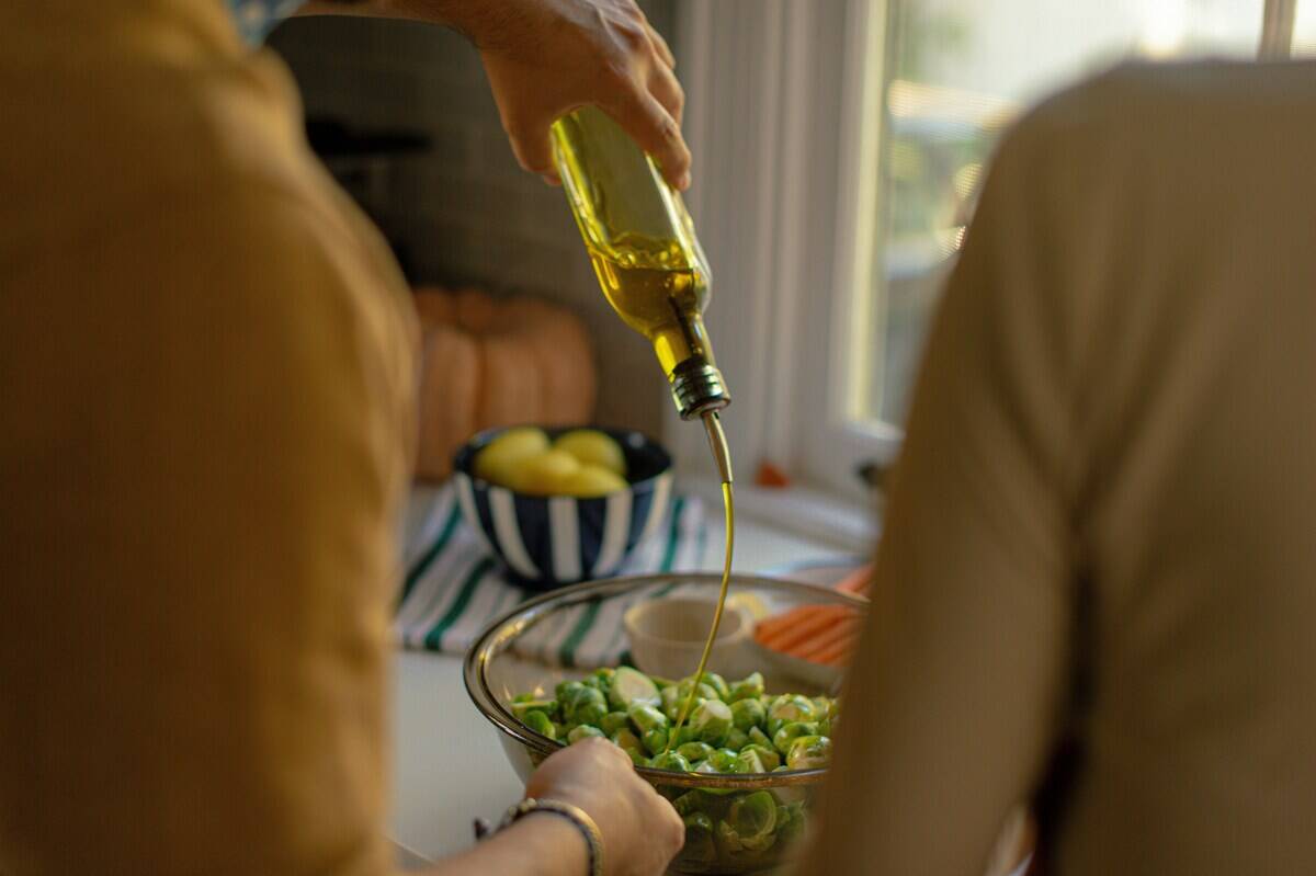 Person pouring olive oil onto vegetables in a glass bowl while another watches