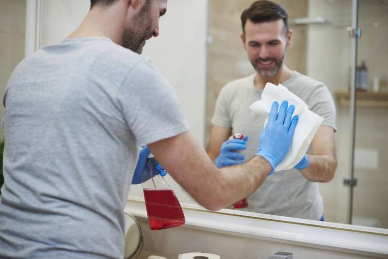 Man cleaning bathroom mirror with paper towel and spray bottle. 