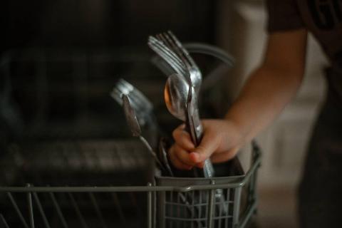 A child placing silverware in a dishwasher with the handles down. 