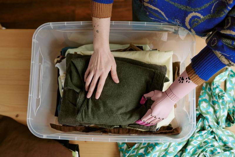 A person with a prosthetic arm putting clothes in a clear plastic bin. 