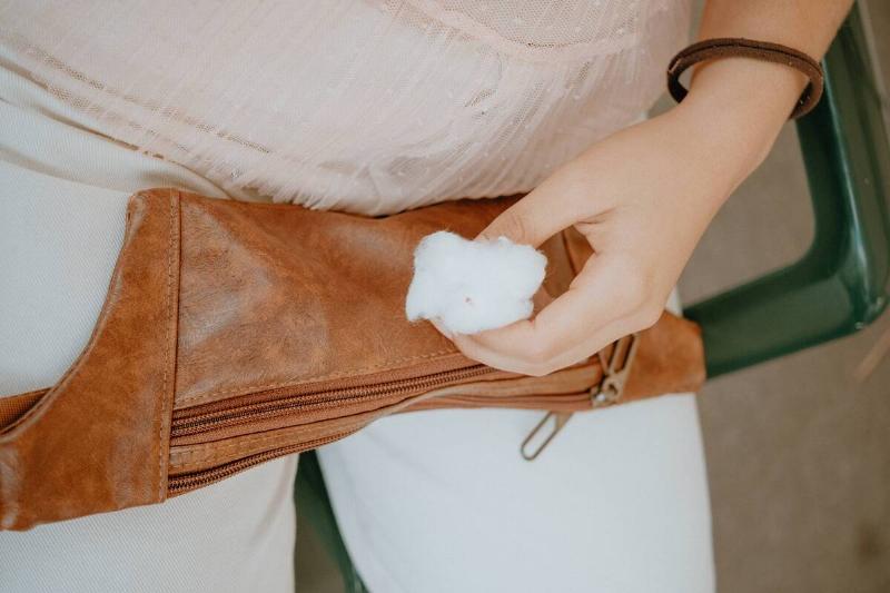 Woman holding a cotton swab near a brown leather bag. 
