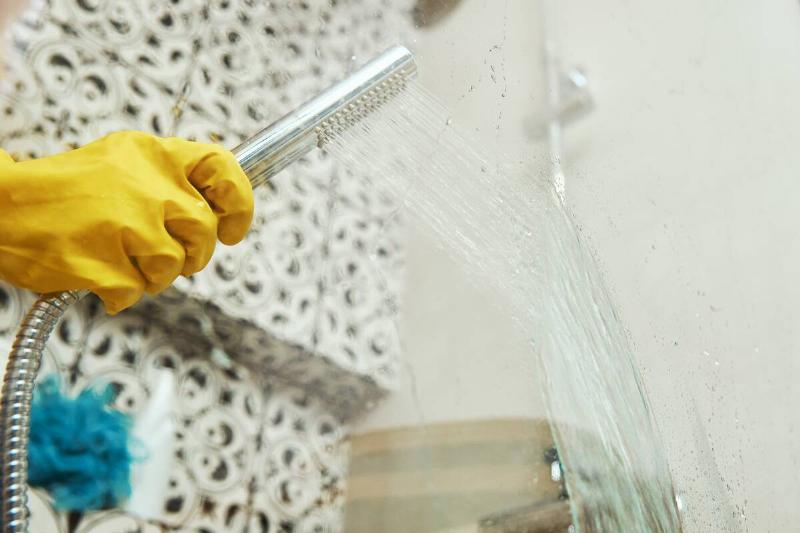 Woman in yellow rubber gloves cleans glass bathroom shower with shower head.