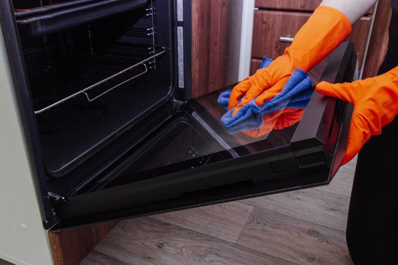 Women's hands in gloves with a microfiber cleaning cloth clean the oven door from the inside.