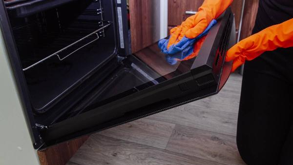 Women's hands in gloves with a microfiber cleaning cloth clean the oven door from the inside.