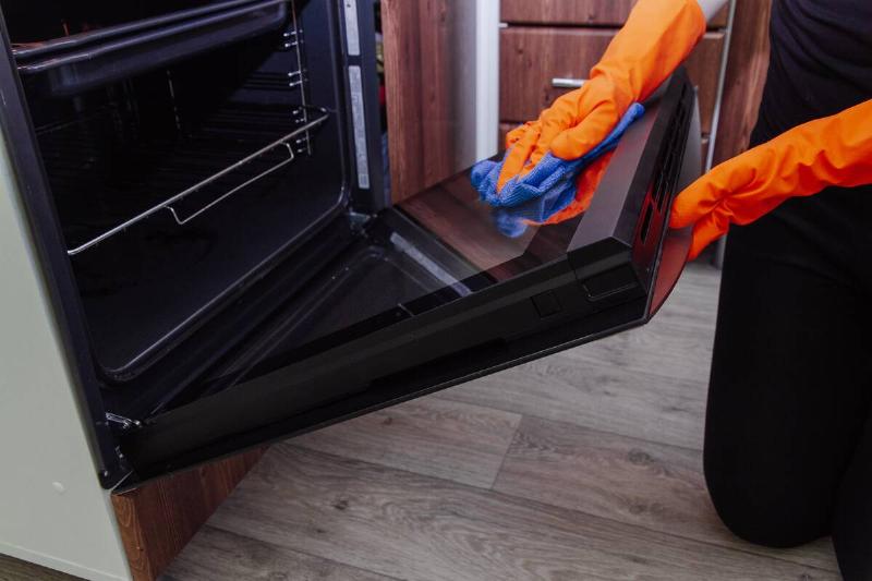 Women's hands in gloves with a microfiber cleaning cloth clean the oven door from the inside.