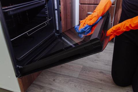 Women's hands in gloves with a microfiber cleaning cloth clean the oven door from the inside.