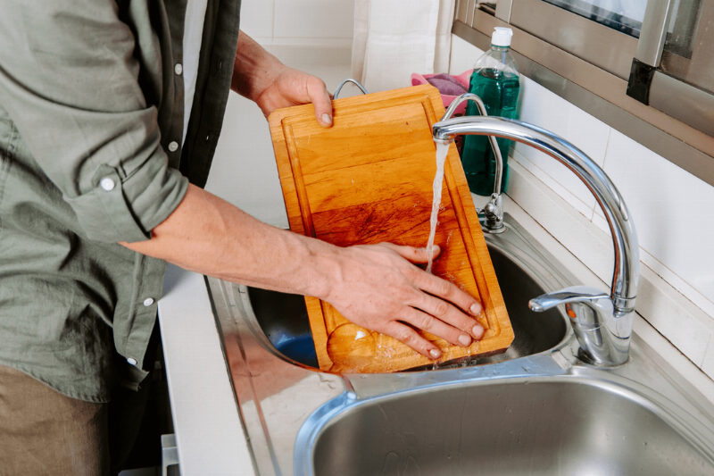 Hand rinsing a wooden cutting board under a sink tap. 