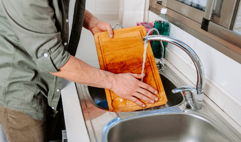 Hand rinsing a wooden cutting board under a sink tap. 