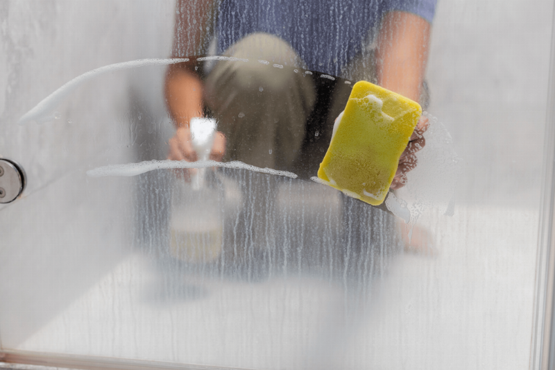 Person cleaning glass shower with sponge and spray bottle. 