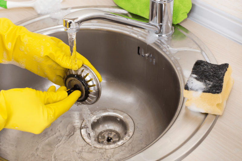 Hands in yellow rubber gloves cleaning out a sink drain.