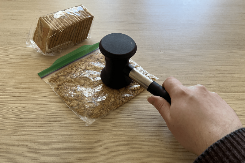 A hand crushing crackers in a plastic bag with a mallet.