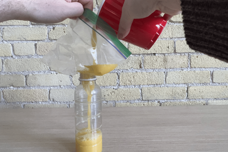 Hand pouring soup into bottle using a ziploc bag funnel.