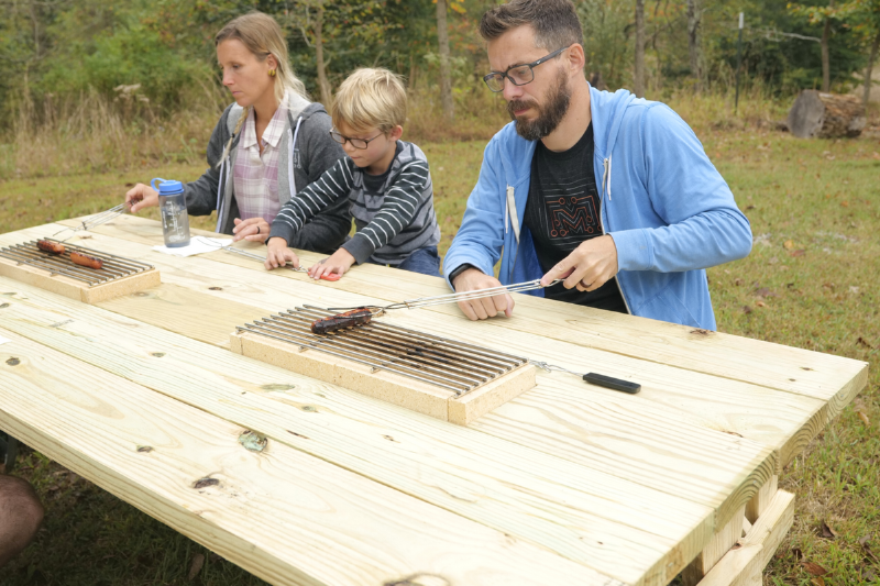 A family grilling hot dogs on a picnic table outside. 