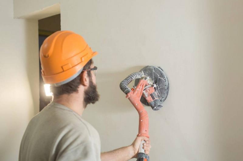 A bearded man sanding a wall. 