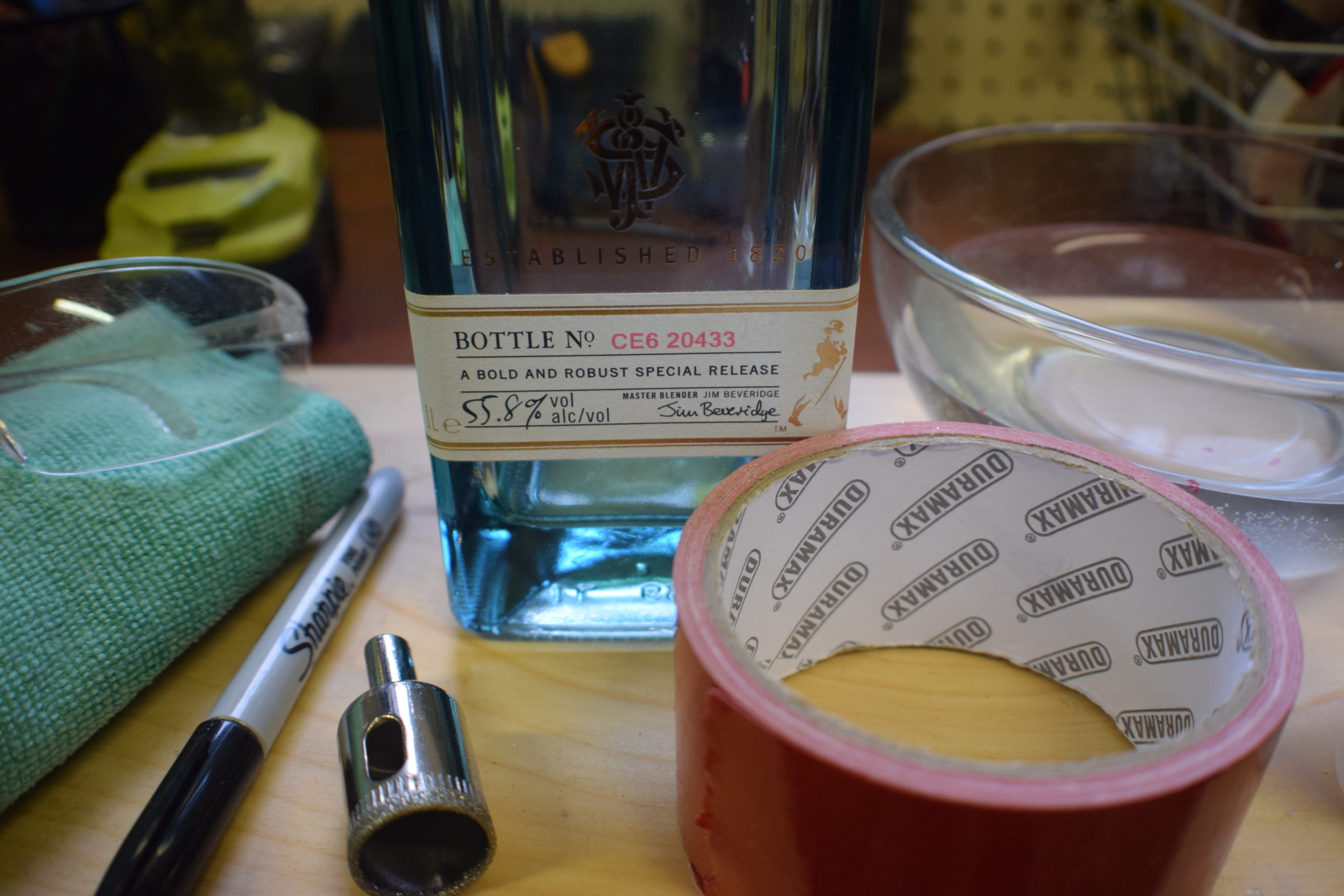 Supplies for making a bottle lamp. Sharpie, diamond drill bit, towel, safety glasses, duct tape, water and a bottle on wood slab.