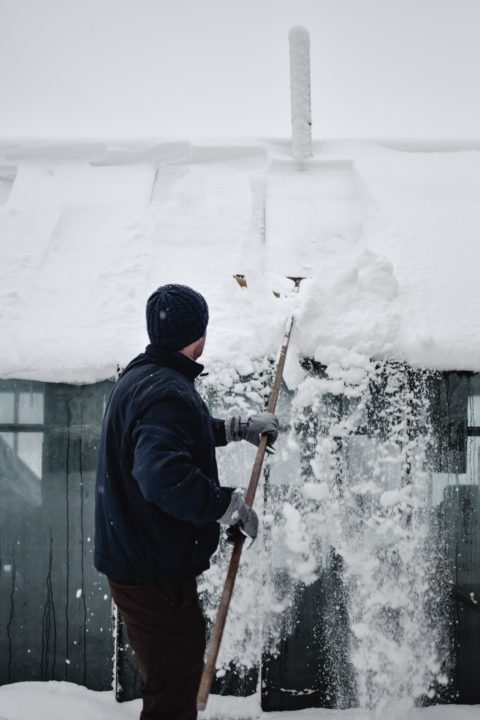 man in black coat and had brushing snow off of a shed roof in the winter