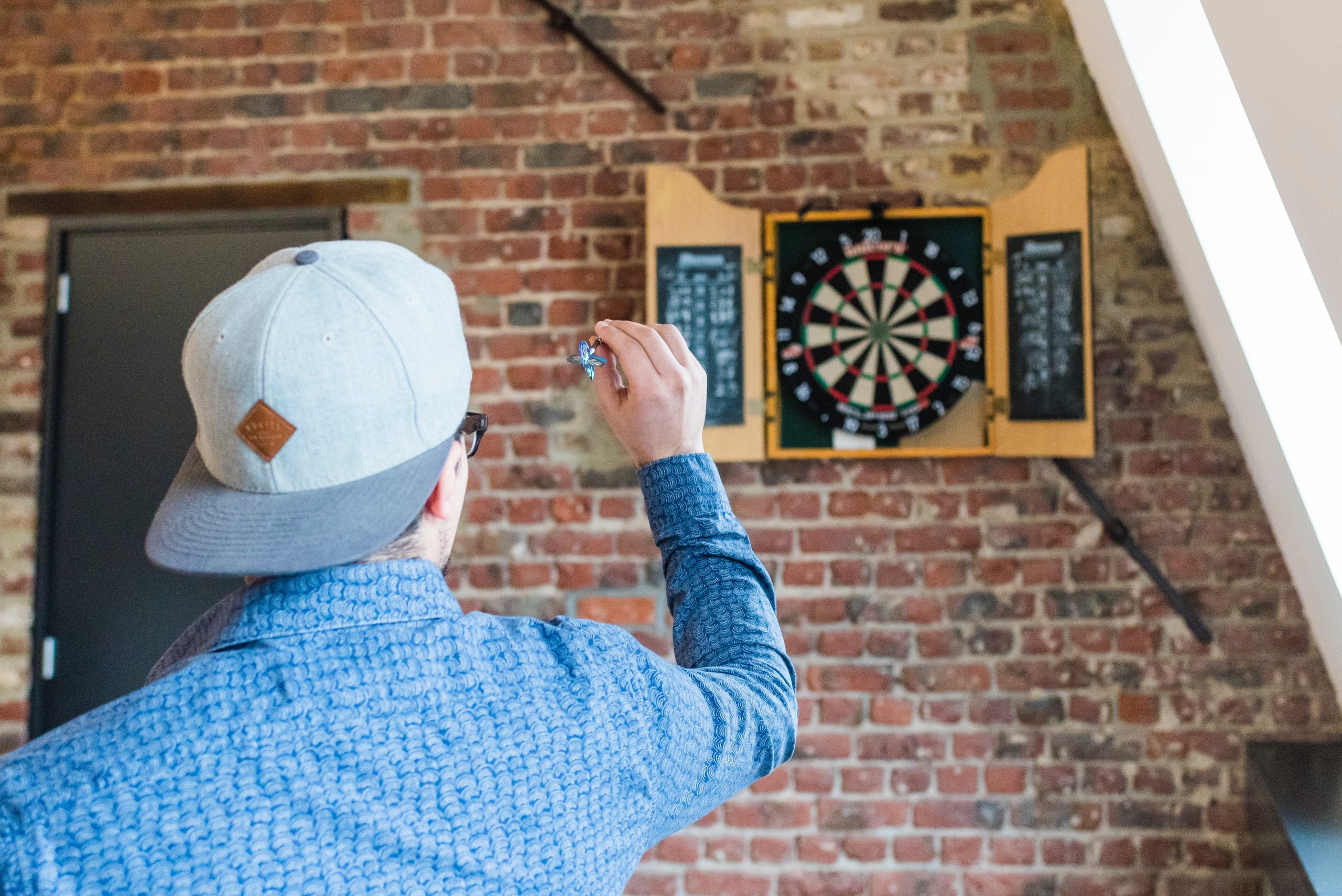 man with hat on backwards and a blue shirt tossing a dart at a dartboard on a brick wall